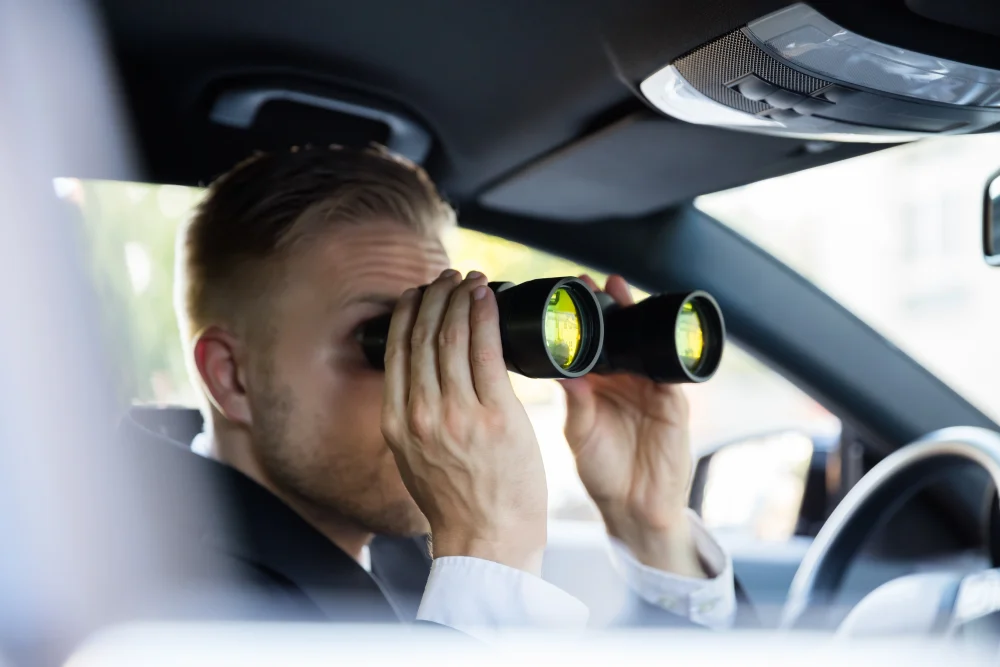 An image showing a man holding black binoculars while sitting in the driver's seat of a car.