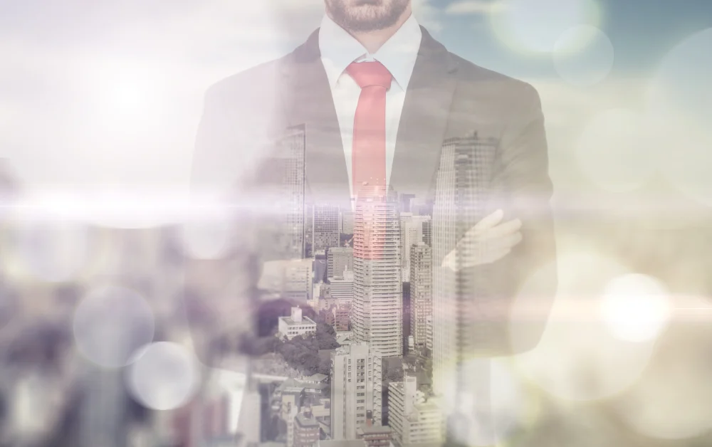 This image shows a businessman wearing a formal suit and red tie, visualized through a double exposure technique that blends the city skyline into the suit