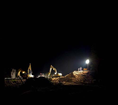 This image depicts a construction site operating at night, illuminated by a single high-mast light tower.